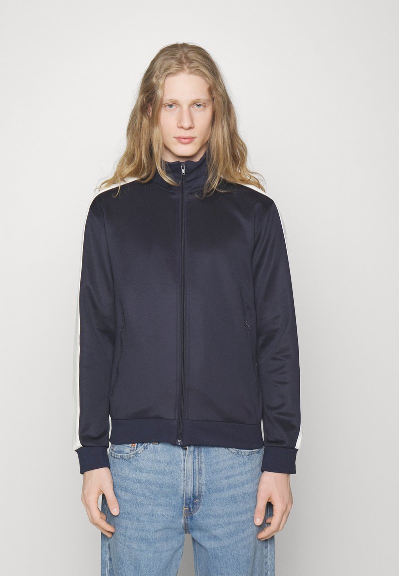 Veste zippée bleu marine en tissu lisse, avec des rayures latérales blanches, un col haut et des poignets et un ourlet côtelés.