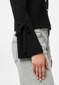 Suéter negro de punto con mangas acampanadas y cierres de lazo. Combinado con jeans de mezclilla gris claro, que muestran un acabado de tejido texturizado.