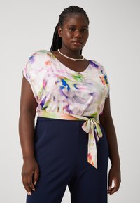 Blouse à motif floral en tissu léger et fluide, avec un décolleté arrondi et des manches courtes, présentant un détail de taille nouée dans des couleurs pastel.