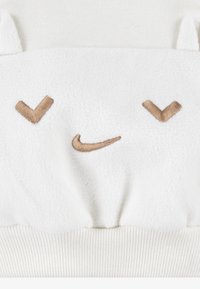 Balts vilnas džemperis ar spilgti priekšpusi, uz kuras ir izšūts smaidīgs sejiņa un brūni akcenti. Riblēta apakšmalas šuve piedod papildu tekstūru.