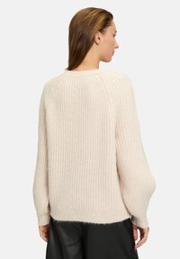 Maglione spesso di colore crema con texture a coste, vestibilità ampia e spalle scese. Maniche e orlo finiti. Indossato sopra pantaloni neri.