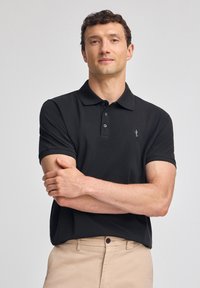 Svart pikétröja i mjukt material. Har en klassisk krage, tre knappar och en liten broderad logotyp på vänster bröst.