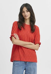Rød øjenløs bluse med korte flæseærmer, der har en tekstureret overflade og diamantmønstre. Kombination med blå denim jeans.