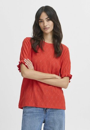 Blouse rouge à œillets avec des manches courtes à volants, dotée d'une surface texturée et de motifs en diamant. Associée à un jean en denim bleu.