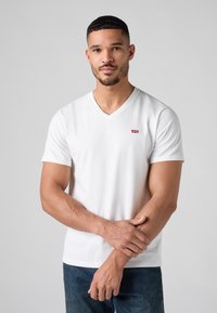 Valkoinen lyhythihainen V-pääntie T-paita, jossa on pieni punainen Levi's-logo vasemmassa rinnassa, valmistettu pehmeästä puuvillakankaasta. Yhdistetty sinisiin farkkuihin.