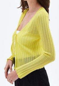 Kobieta w prążkowanym, prześwitującym żółtym kardiganie zawiązanym z przodu na białą bluzkę, w czarnych spodniach na białym tle.