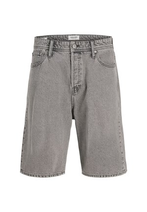 Niño de pie y sonriendo, con una camiseta beige de punto de manga corta, pantalones cortos grises hasta la rodilla, calcetines blancos y zapatos negros sobre un fondo liso.