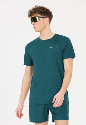 Young man wearing mirrored sports sunglasses, a teal "Endurance" T-shirt, and matching shorts, looking to the side against a white background.