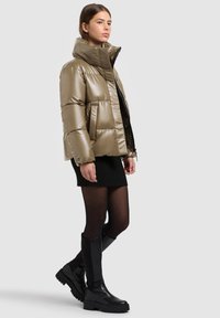 Doudoune vert olive avec un col montant, une finition brillante et un design matelassé ; associée à une jupe noire tricotée et des bottes noires montantes jusqu'aux genoux.