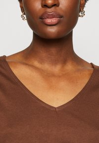 Brown V-neck top with a smooth texture, featuring a subtle seam detailing at neck and delicate gold hoop earrings with a wave design.