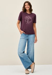 Camiseta de manga corta morada con un diseño impreso, combinada con pantalones vaqueros de pierna ancha de color azul claro y zapatos beige. Material de algodón suave.