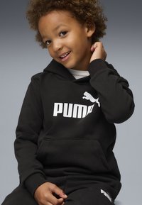 Ensemble de sweat à capuche noir et de pantalon de survêtement en matériau doux, avec un logo Puma blanc bien visible sur le devant et la jambe gauche du pantalon.