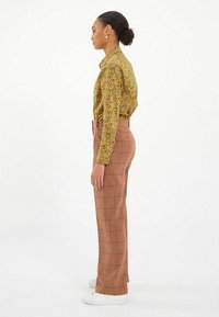 Chemise jaune à motifs floraux avec des manches longues, associée à un pantalon à carreaux taille haute rouge et marron, portée avec des baskets blanches.