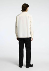 Camicia button-up di un leggero crema con una vestibilità rilassata, maniche lunghe e pantaloni neri, vista da dietro su uno sfondo neutro.