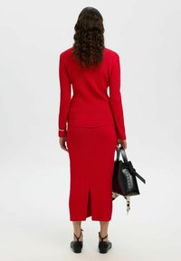 Robe en tricot à côtes rouges en deux pièces avec des manches longues et une jupe à ourlet fendu, accompagnée d'un sac à main noir avec un accent en nœud.
