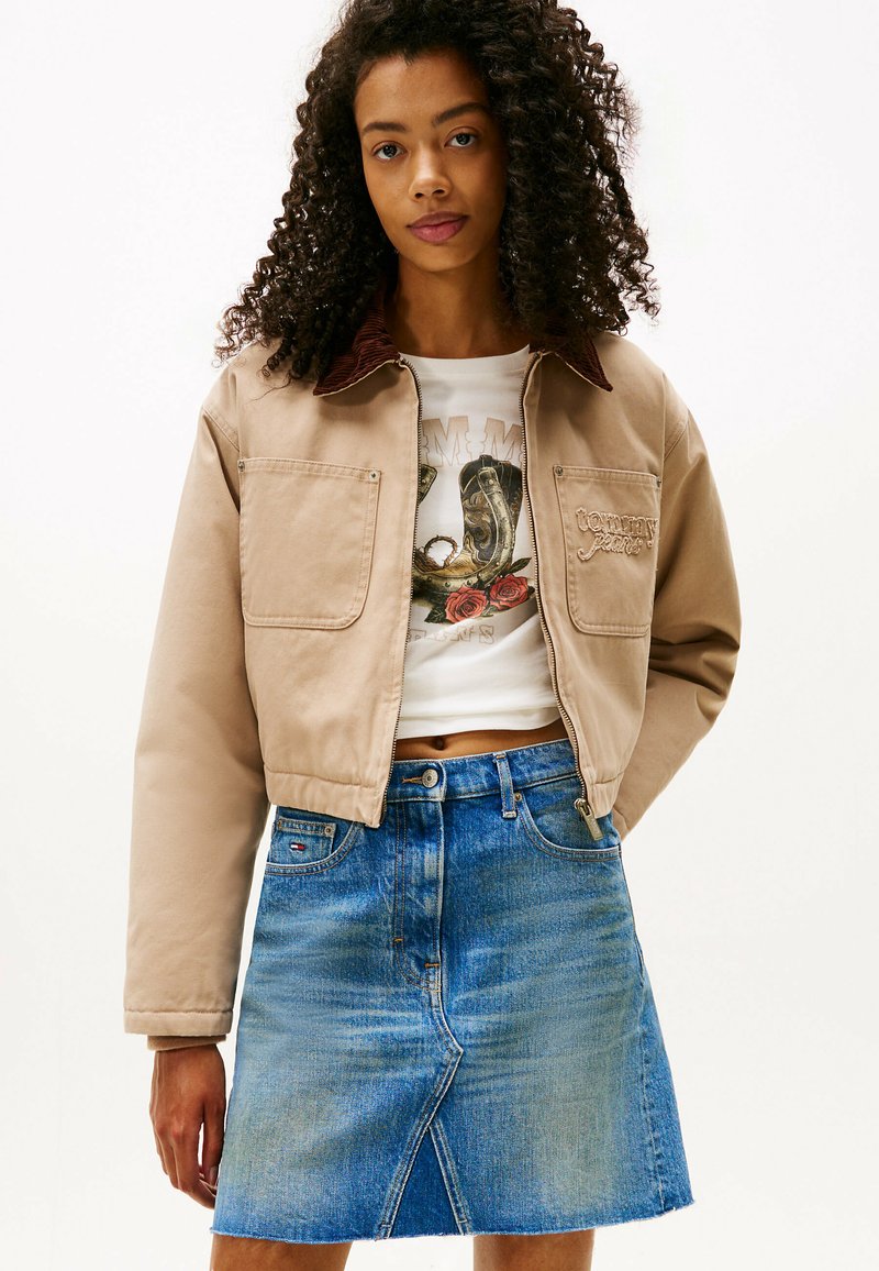 Chaqueta beige con cuello de tergal marrón, con dos bolsillos en el pecho, combinada con una camisa gráfica blanca ajustada y una falda de mezclilla en forma de A.