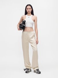 Femme debout portant un crop top blanc, un pantalon ample beige, des baskets noires et blanches, tenant un sac à main noir sur fond uni.