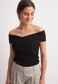 Top nero a spalla scoperta in tessuto a coste con design attorcigliato sul seno; abbinato a pantaloni grigi chiari a vita alta. Texture liscia in tutto il capo.