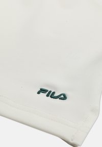 Valkoinen kangas, jossa vihreä FILA-logo brodeerattuna ommellun reunuksen lähellä.