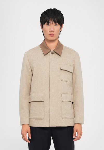 Giacca in misto lana beige con colletto marrone, chiusura frontale con bottoni e quattro tasche utili. Design semplice e strutturato con una superficie liscia.