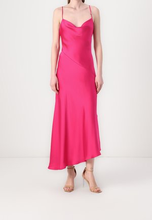 Kvinde iført en ærmeløs, knaldrød satin midi-kjole med tynde stropper og beige åbne højhælede sandaler, står mod en enkel baggrund.