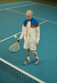 Conjunto deportivo con una chaqueta de cremallera en crema y azul, pantalones cortos crema y zapatillas grises. Sosteniendo una raqueta de tenis naranja, preparándose para golpear una pelota.