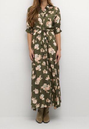 Kvinde iført en olivengrøn lang kjole med blomsterprint, knapper og et bælte bundet i taljen, kombineret med beige ankelstøvler.