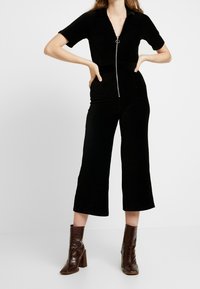 Jumpsuit de pana negra con mangas cortas, escote en V, cremallera frontal y piernas anchas recortadas. Combinado con botines de tacón marrones.