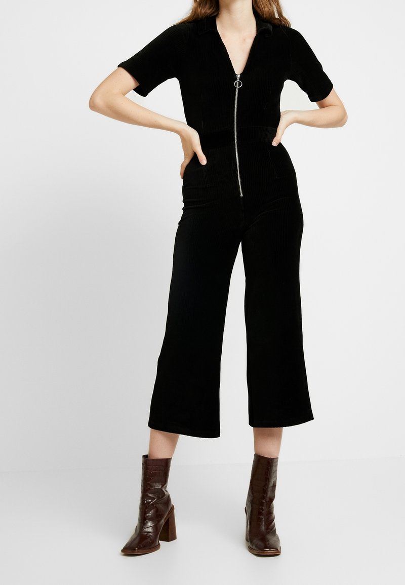 Jumpsuit de pana negra con mangas cortas, escote en V, cremallera frontal y piernas anchas recortadas. Combinado con botines de tacón marrones.