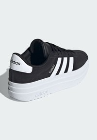 Baskets en suède noir avec semelle en caoutchouc blanc, trois bandes blanches sur le côté, et un logo sur le talon et la languette.