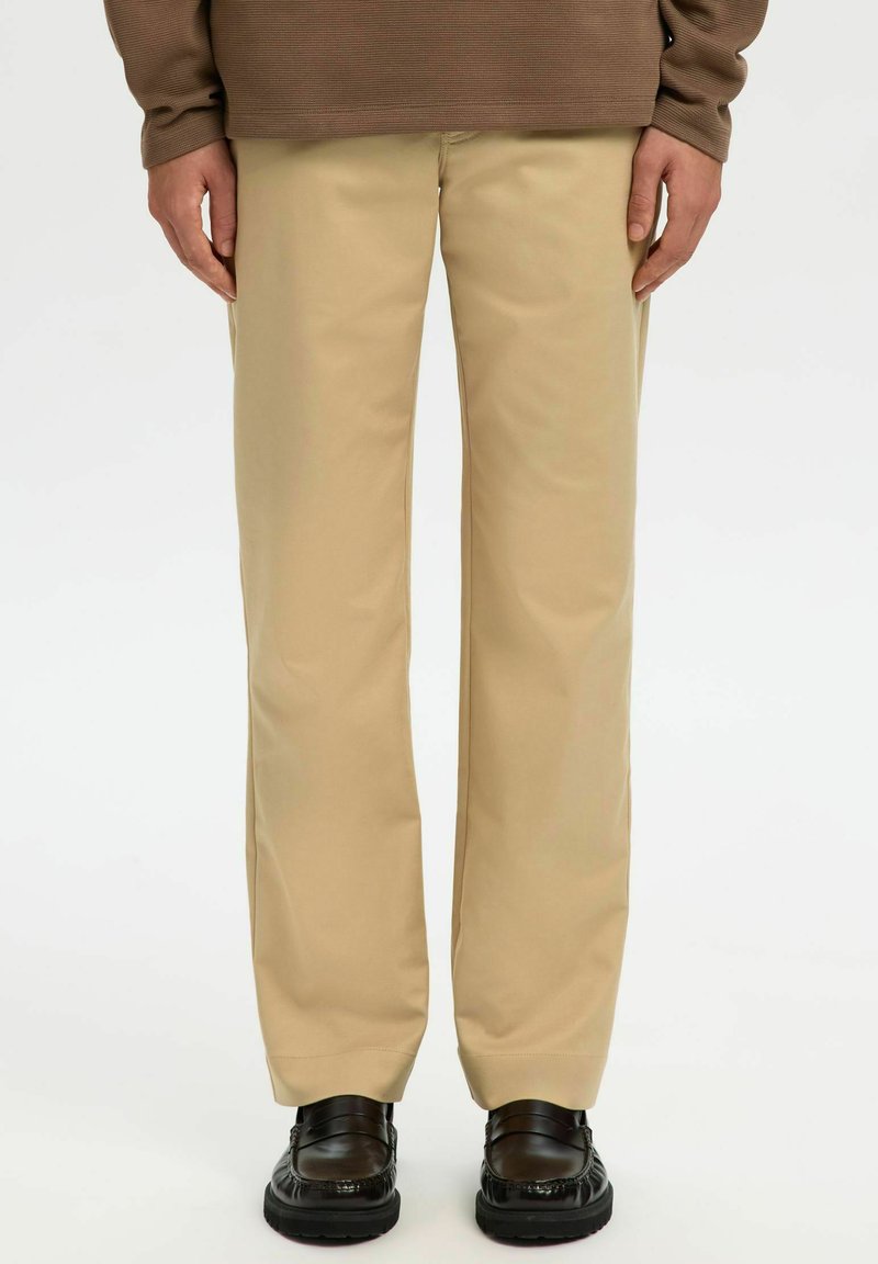 Bež hlače s krojem naravnost navzdol iz gladkega blaga. Imajo subtilne detajle šivov. Nošene z temnimi, sijočimi čevlji.