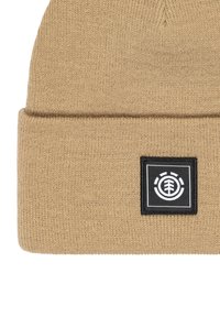 Gorro de punto beige con un dobladillo. Presenta un parche negro cuadrado con un logotipo blanco. El material parece suave y texturizado.