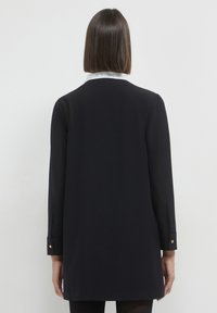 Lang�ärmlige schwarze Tunika, glattes Material, klassischer Kragen mit weißem Saum, goldene Knopfakzente an den Manschetten, gerade Silhouette.