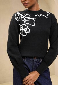 Maglione nero lavorato a maglia con maniche lunghe, caratterizzato da ricami floreali bianchi e un collo a coste. Indossato con jeans di denim scuro.