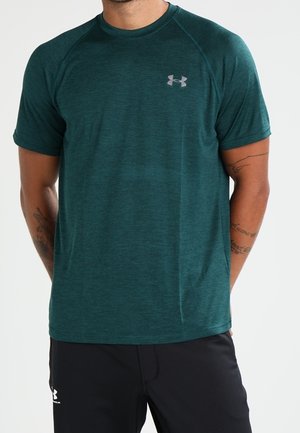 Mężczyzna ubrany w turkusową sportową koszulkę Under Armour z krótkim rękawem oraz czarne spodenki Under Armour, stojący z rękami za plecami.