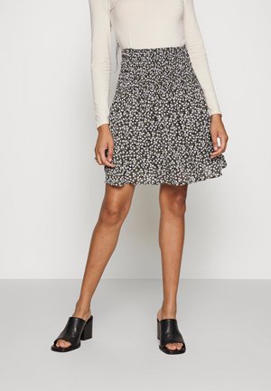 Femme portant une jupe noire fleurie, un haut beige à manches longues et des chaussures noires à talons blocs ouvertes à l'avant, debout devant un fond uni.