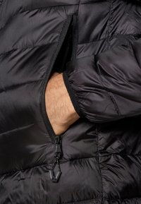 Casaco puffer preto com um bolso com fecho. O tecido texturizado apresenta um padrão acolchoado e punhos elásticos. O fecho tem uma pala de puxar preta.