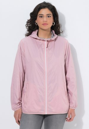 Vrouw met donker krullend haar, zilveren oorringen in de vorm van hoops, een lichtroze capuchonvest met ritssluiting en een grijze broek, staand tegen een witte achtergrond.