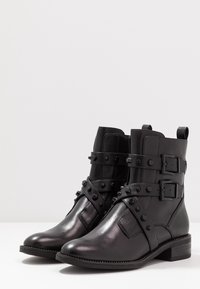 Botas de tobillo de cuero negro con dos correas ajustables, detalles de tachuelas, puntera redondeada y un tacón bajo tipo bloque. Textura suave con acabado pulido.