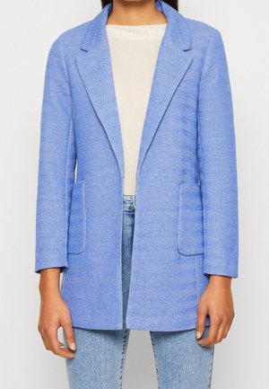 Femme portant un blazer bleu clair à chevrons avec des revers crantés par-dessus un haut blanc et un jean bleu clair, debout devant un fond uni.