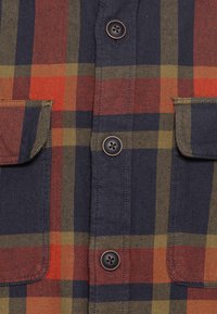 Plaid button-up shirt featuring dark navy, red, orange, and olive green colors with two chest pockets and brown buttons. Cotton fabric.