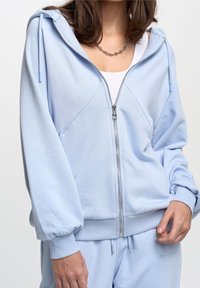 Light blue zip-up hoodie with a relaxed fit, drawstring hood, and ribbed cuffs. Soft fabric features a subtle texture and sporty design.