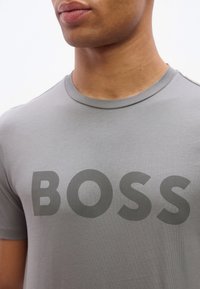 Άνδρας που φοράει ανοιχτό γκρι μπλουζάκι με στρογγυλή λαιμόκοψη, με τη λέξη "BOSS" τυπωμένη με μεγάλα σκούρα γκρι γράμματα στο στήθος.