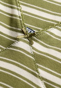 Tissu côtelé rayé vert et blanc avec des surpiqûres décoratives et un petit nœud vert olive près d'une étiquette de vêtement partiellement visible.