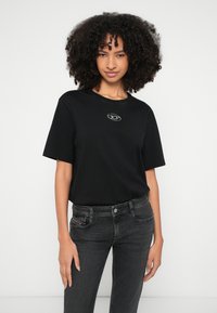 Γυναίκα με σγουρά μαύρα μαλλιά, φοράει μαύρο T-shirt με μικρό λευκό λογότυπο και σκούρα γκρι τζιν, στέκεται μπροστά σε απλό λευκό φόντο.