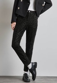 Pantalons à imprimé léopard noir en tissu doux, avec une coupe slim, des coutures latérales et une taille haute, associés à des chaussures à lacets noires.