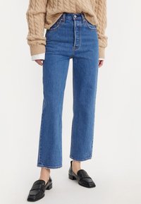 Personne portant un jean bleu droit taille haute, un pull en maille torsadée beige sur une chemise blanche et des mocassins noirs sans lacets.