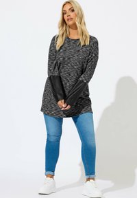 Schwarz-gestreifte Tunika mit grauen Streifen und schwarzen Kunstlederärmeln, kombiniert mit blauen Skinny-Jeans und weißen Sneakers. Glatte Textur, lässiges Design.