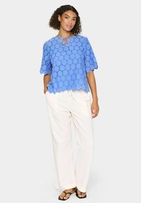 Top in pizzo azzurro chiaro con bordi scallopati e maniche corte, caratterizzato da un colletto a oblò; abbinato a pantaloni bianchi a gamba larga e infradito nere.