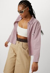 Hellblau gemustertes Hemd über einem weißen Croptop, kombiniert mit khakifarbenen Shorts mit einem gemusterten Bund. Accessoires umfassen Sonnenbrille.